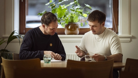 To mænd sidder ved et træbord i en lys café, mens de samtaler og gennemgår noter, omkranset af grønne planter i vindueskarme.